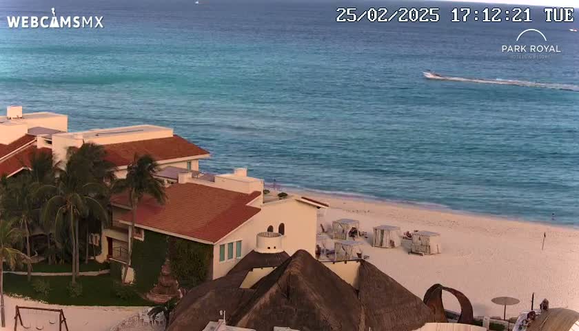 A beachfront resort with red-tiled roofs and palm trees overlooks a calm turquoise ocean and a sandy beach under a clear sky.