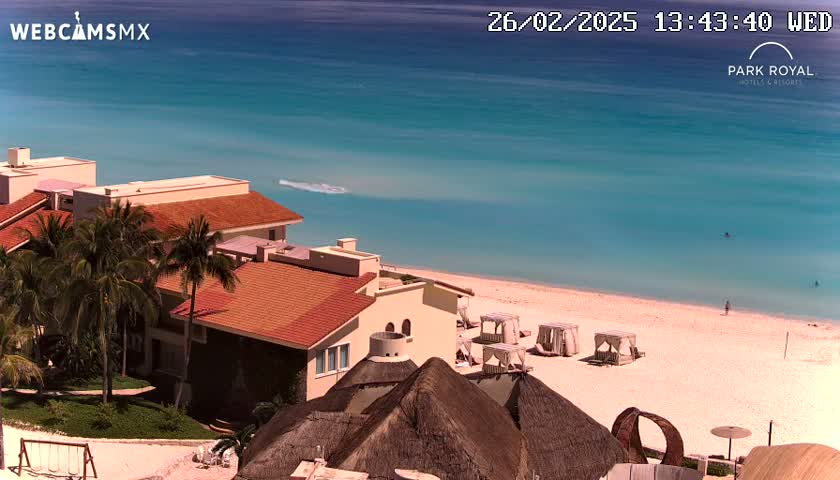 An aerial view shows a resort with red-tiled buildings, palm trees, and thatched-roof structures overlooking a calm, turquoise ocean and a sandy beach on a sunny day.