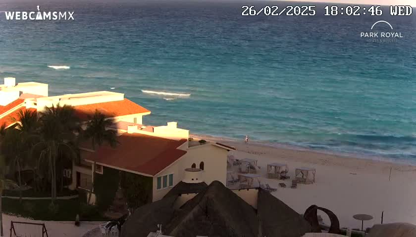 An ocean beach with teal-colored water and small waves at sunset, is seen from above a resort with red-roofed buildings and palm trees.