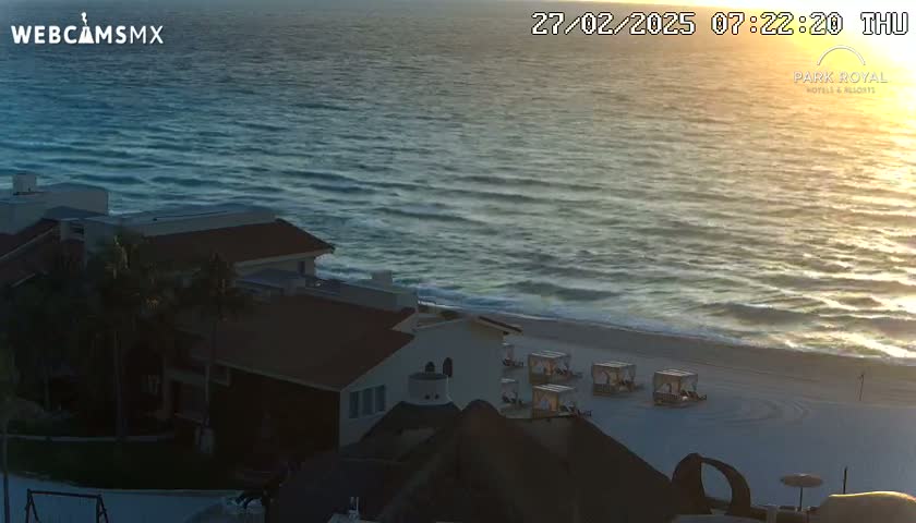 A beachfront building is seen at sunrise, with calm ocean waves lapping a sandy shore.