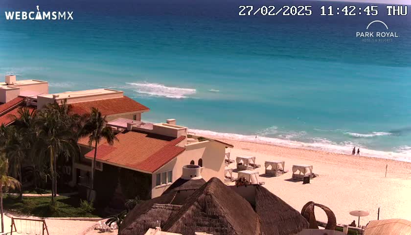 A resort with red-tiled roofs and palm trees overlooks a beach with turquoise water and gentle waves under a sunny sky.