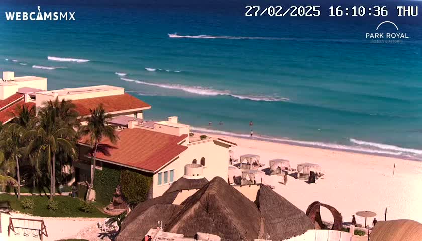 An aerial view shows a beachfront resort with red-tiled buildings and thatched-roof structures, overlooking a sandy beach with gentle waves and turquoise water on a sunny day.