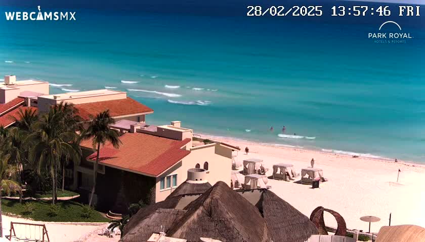 A sunny day shows a beach with turquoise water, small waves, and several beach cabanas, next to a resort with red-tiled roofs and palm trees.