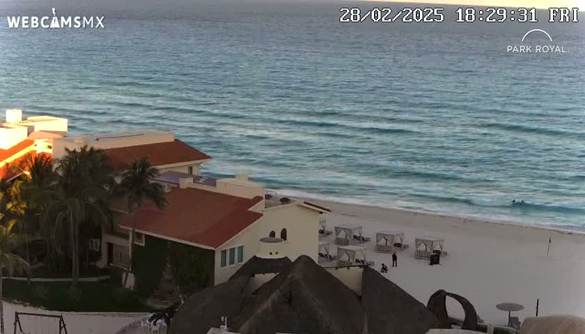 A beachfront hotel with palm trees is situated above a sandy beach with calm ocean waves under a twilight sky.