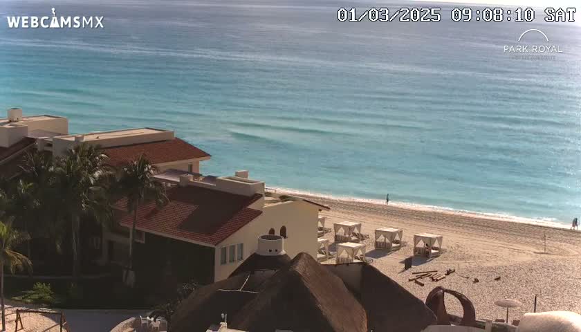 An oceanfront resort with red-tiled roofs and palm trees overlooks a calm, turquoise ocean and sandy beach on a sunny day.