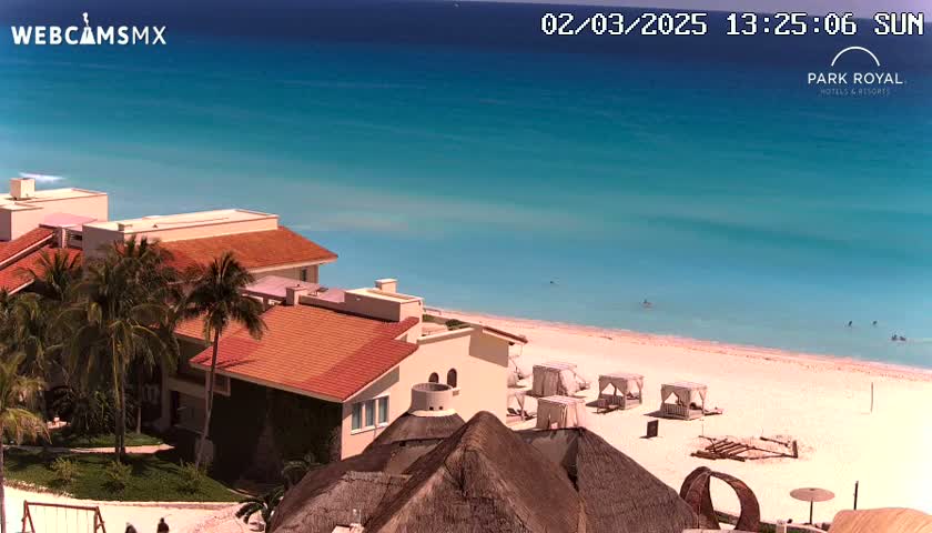 A sunny day shows a beachfront resort with terracotta-roofed buildings, palm trees, and beach umbrellas on a white sand beach next to turquoise water.