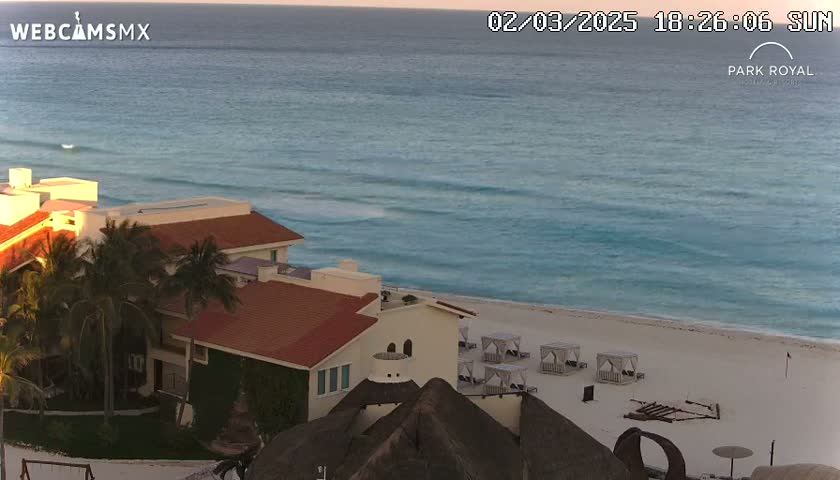 A beachfront resort with red-tiled roofs and palm trees overlooks a calm turquoise ocean under a clear sunset sky.
