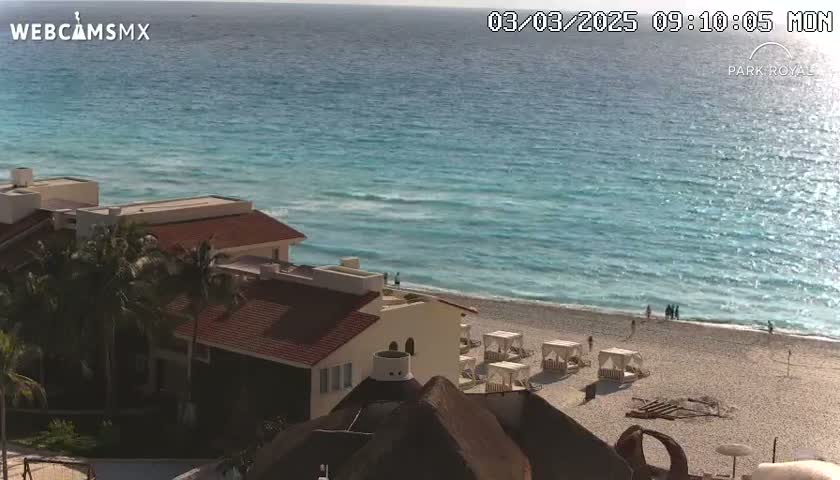 A sunny day at a beach resort shows a turquoise ocean with small waves lapping a sandy shore where several beach cabanas are set up in front of a building with a red tile roof.