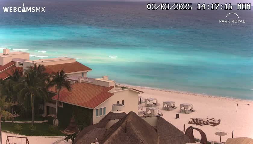 A resort with red-tiled roofs sits on a white-sand beach overlooking a calm, turquoise ocean.