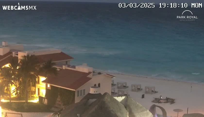 A beachfront resort at dusk shows a calm ocean and a sandy beach with some beach furniture.