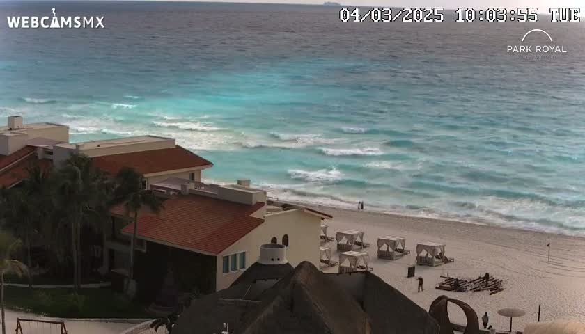 A beachfront resort with red-tiled roofs overlooks a turquoise ocean with moderate waves under a partly cloudy sky.