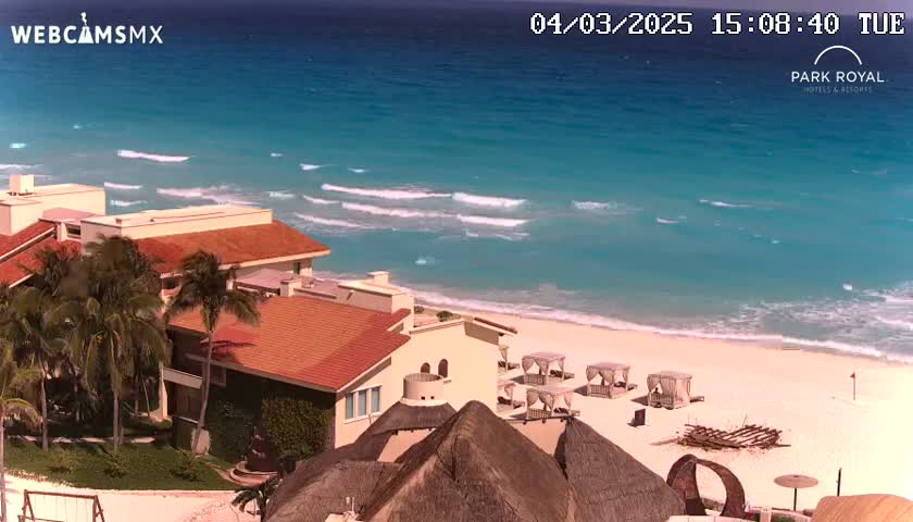 A beachfront resort with red-tiled roofs and palm trees overlooks a calm turquoise ocean with small waves on a sunny day.