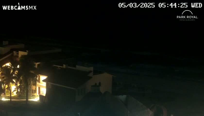 At night, a brightly lit resort building is situated near a dark ocean under a clear sky.