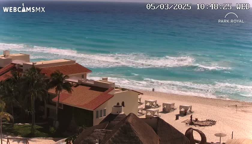 A beachside resort with red-tiled roofs overlooks a bright turquoise ocean with moderate waves on a sunny day.