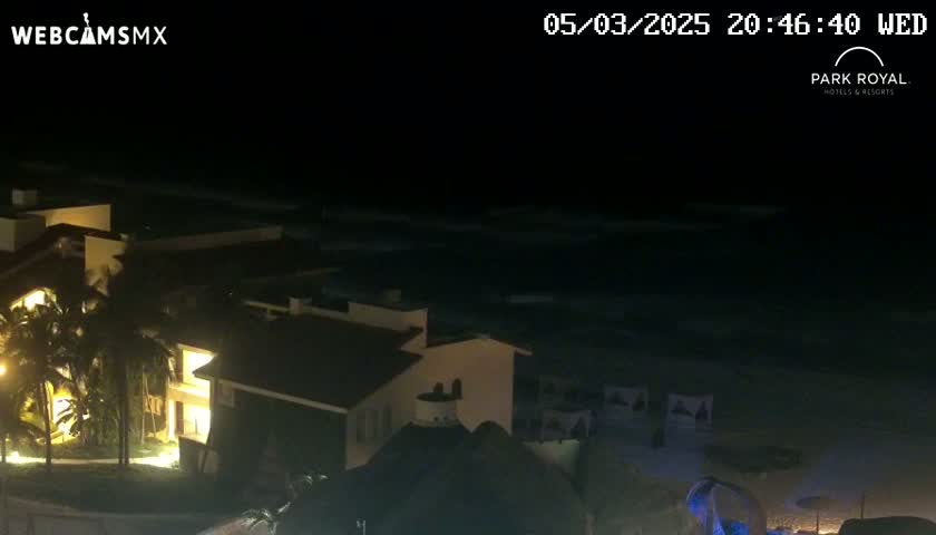 At night, a beachside resort is illuminated, overlooking a dark ocean with gentle waves.