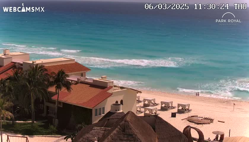 A beachfront resort with red-tiled roofs and palm trees overlooks a tranquil turquoise ocean with gentle waves on a sunny day.