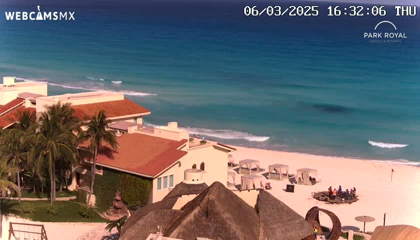 A beachfront resort with terracotta-roofed buildings and palm trees overlooks a calm turquoise ocean with gentle waves on a sunny day.