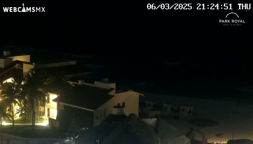A nighttime view of a brightly lit resort complex situated near a dark ocean under a clear, moonless sky.