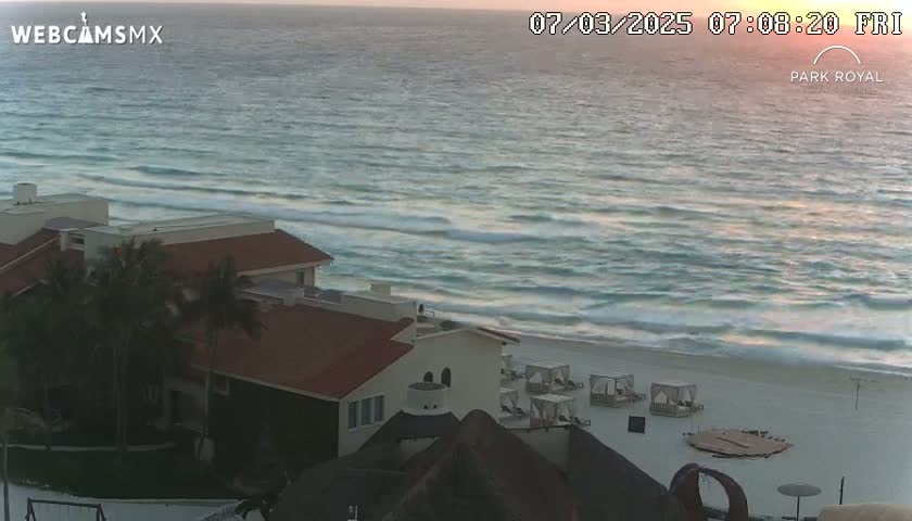 A beachside resort with palm trees overlooks a calm ocean with small waves under a sunrise sky.