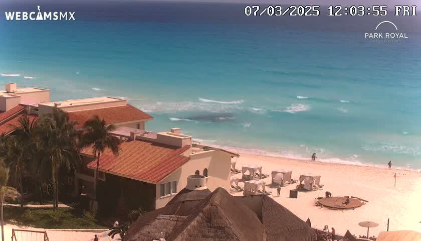 A beachfront resort with red-tiled buildings and palm trees overlooks a clear turquoise ocean with gentle waves on a sunny day.