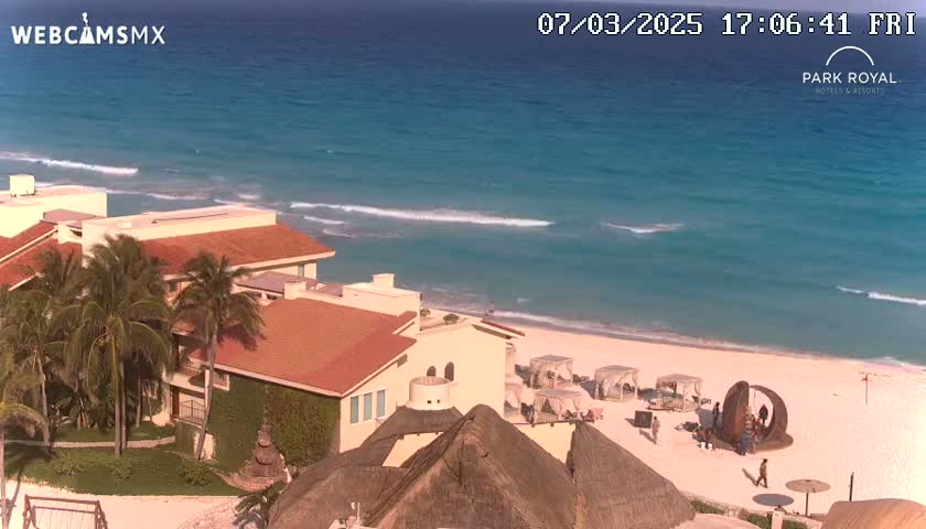 A beachfront resort with red-tiled roofs and palm trees overlooks a sandy beach and turquoise ocean with gentle waves under a sunny sky.