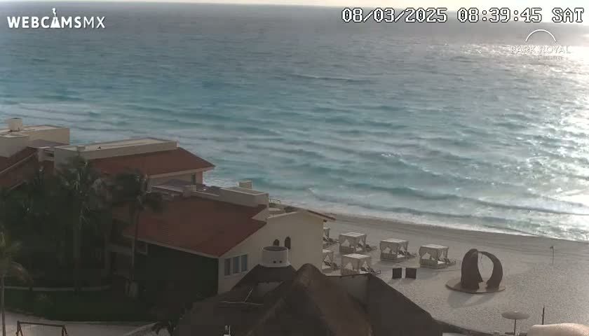 A beachfront resort with red-tiled roofs sits beside a calm, turquoise ocean with small waves under a mostly clear sky.