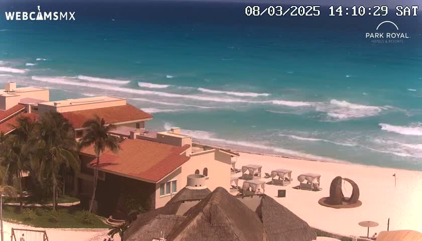 A beachfront resort with red-tiled roofs and palm trees overlooks a sandy beach with gentle waves on a sunny day.