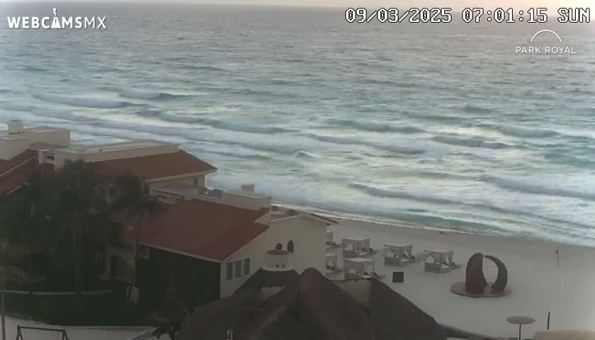 A beachfront resort with red-tiled roofs overlooks a beach with moderate waves under a mostly cloudy sky.