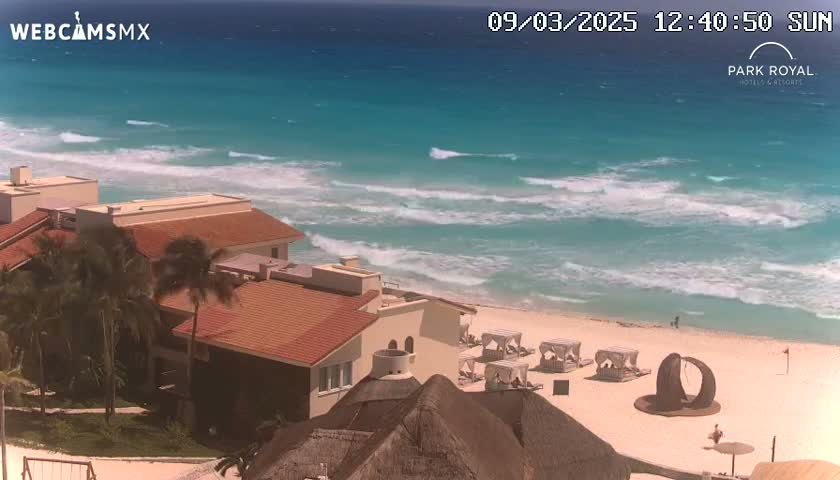 A beach with turquoise water and moderate waves is viewed from above, showing a resort with red-tiled roofs and palm trees.