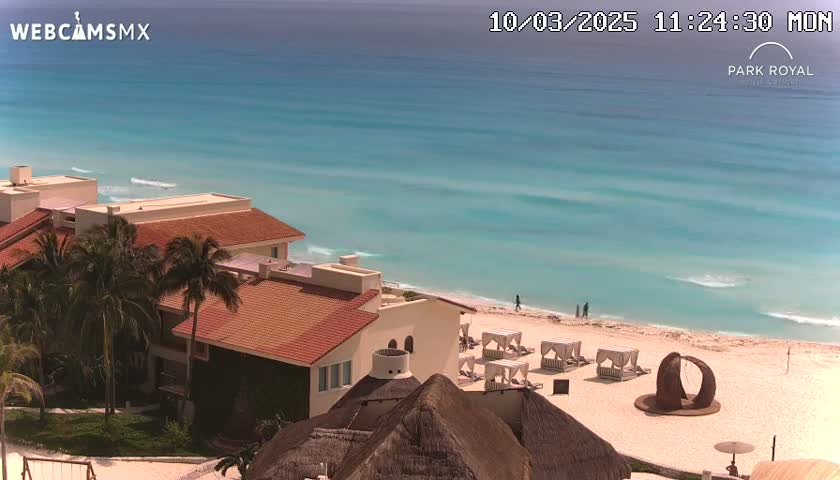 A beachfront resort with red-tiled roofs and palm trees overlooks a calm, turquoise ocean on a sunny day.