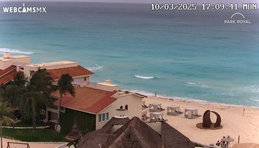 A resort with red-tiled roofs sits on a beach overlooking a turquoise ocean with small waves under an overcast sky.