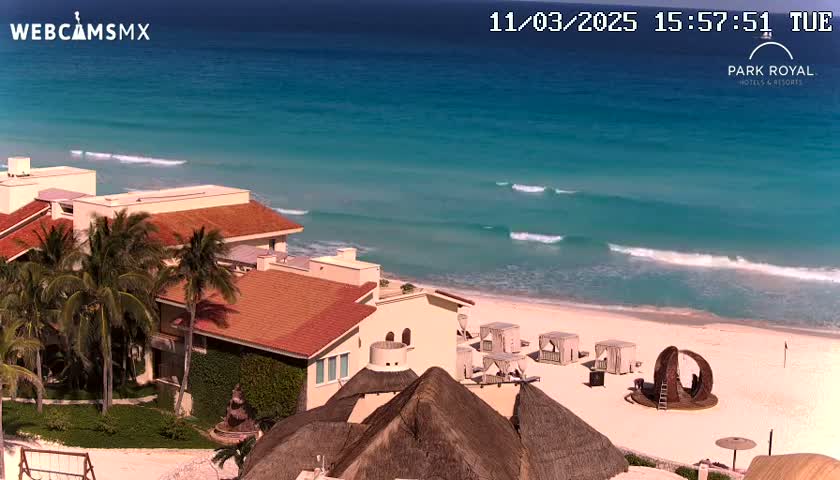 A beachfront resort with red-tiled roofs and palm trees overlooks a clear turquoise ocean with gentle waves under a sunny sky.