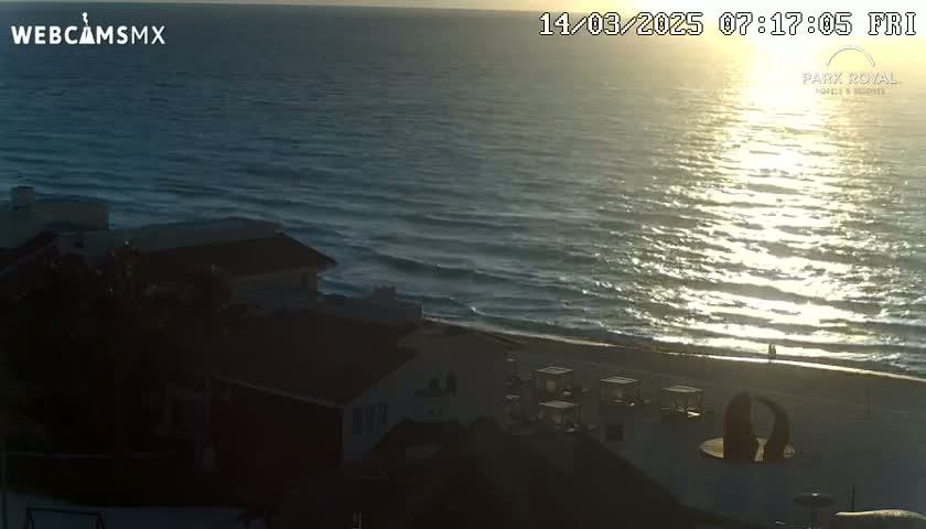 A sunrise illuminates the ocean waves as they lap a sandy beach near a dark building.