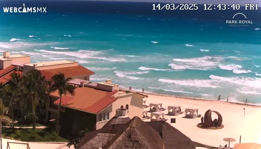A beachfront resort with red-tiled roofs sits beside a turquoise ocean with moderate waves on a sunny day.