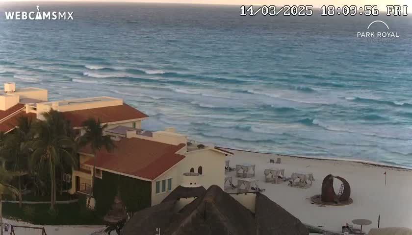 A beachfront resort with reddish-brown roofs and tropical landscaping overlooks a turquoise ocean with gentle waves under a mostly clear sky.