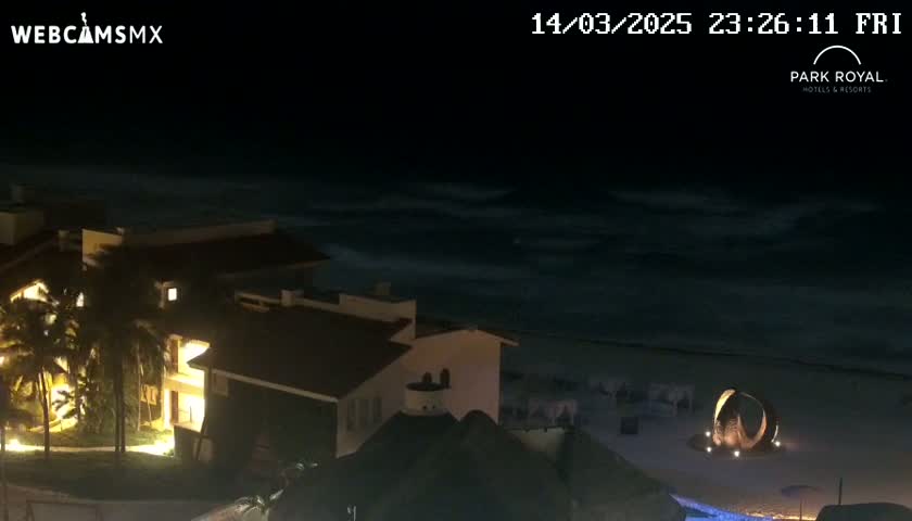At night, a beachfront resort is illuminated, overlooking a dark ocean with moderate waves.