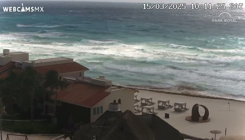 A beachfront resort with red-tiled roofs overlooks a turbulent ocean with heavy waves under overcast skies.