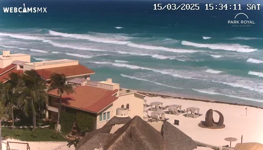 A beachfront resort with red-tiled roofs and palm trees overlooks a sandy beach with moderate waves on a sunny day.