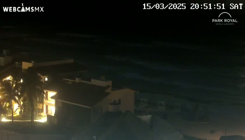 A nighttime view shows a brightly lit building complex near a dark ocean with some small waves under a clear sky.