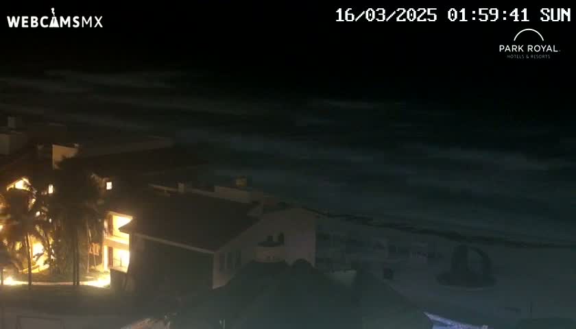 A nighttime, seaside view shows a building with illuminated windows overlooking a dark ocean with subtle wave motion under a clear, dark sky.