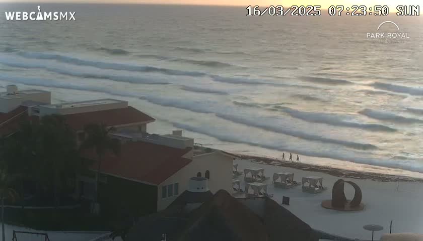 A beach with moderate waves at sunrise, showing several buildings and beach cabanas on the sand.
