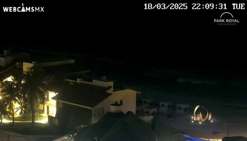 A nighttime, seaside view of a resort with illuminated buildings and palm trees, under a dark sky with a somewhat wavy ocean.