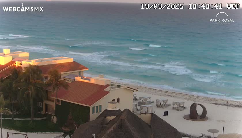 A beachfront resort with red tile roofs and palm trees is visible, overlooking a calm ocean with small waves under a partly cloudy sky.