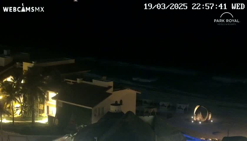 A nighttime view shows a beach with gently lapping waves, illuminated buildings, and palm trees.