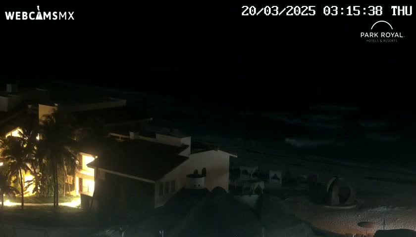 A nighttime view of a brightly lit building complex next to a dark ocean with gentle waves under a clear, dark sky.