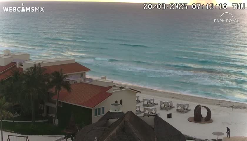 A beachfront resort with red-tiled buildings is shown at sunrise, overlooking a beach with gentle waves and several beach cabanas.