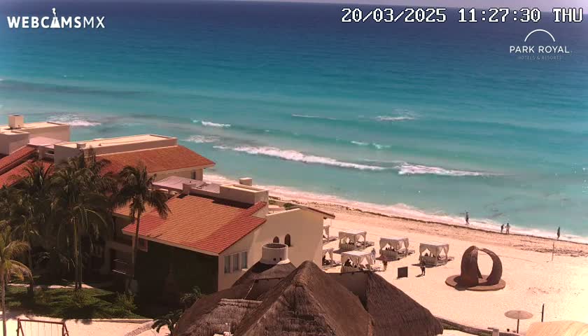 A beachfront resort with red tile roofs and palm trees is shown on a sunny day with turquoise water and small waves gently lapping a sandy shore.