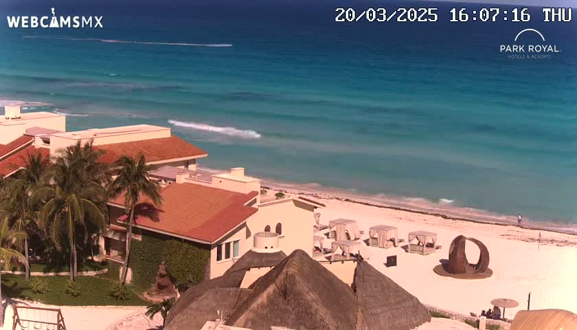 A beachfront resort with red-roofed buildings and palm trees overlooks a calm, turquoise ocean on a sunny day.