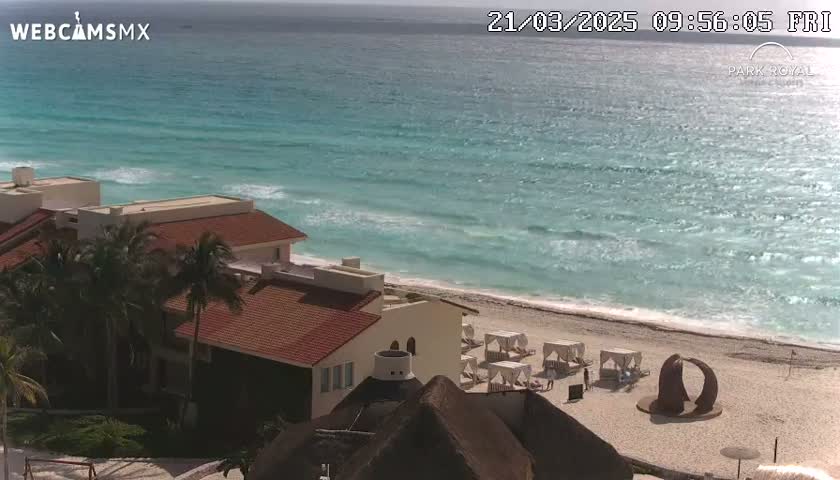 A beachfront resort with red-tiled roofs and palm trees overlooks a sandy beach and turquoise ocean with small waves on a sunny day.