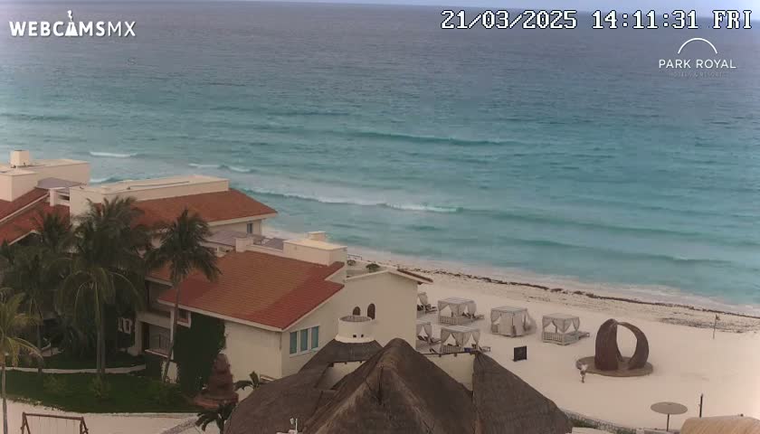 A beachfront resort with red-tiled roofs and palm trees overlooks a turquoise ocean with small waves on a partly cloudy day.